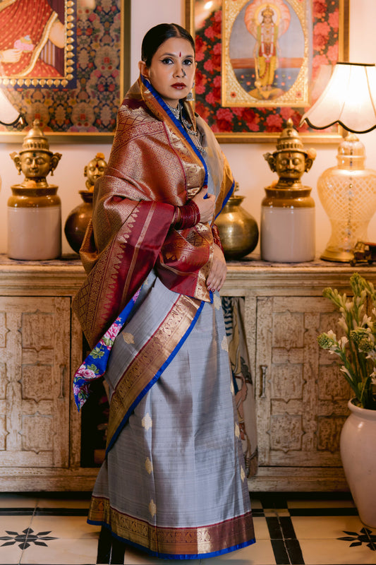 Woman in traditional beautiful handwoven English grey and antique gold Kanjivaram silk saree standing in a decorated room with lamps and artwork.