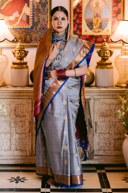 Woman in a traditional beautiful handwoven English grey and antique gold Kanjivaram silk saree standing in a decorated room with lamps and artwork.