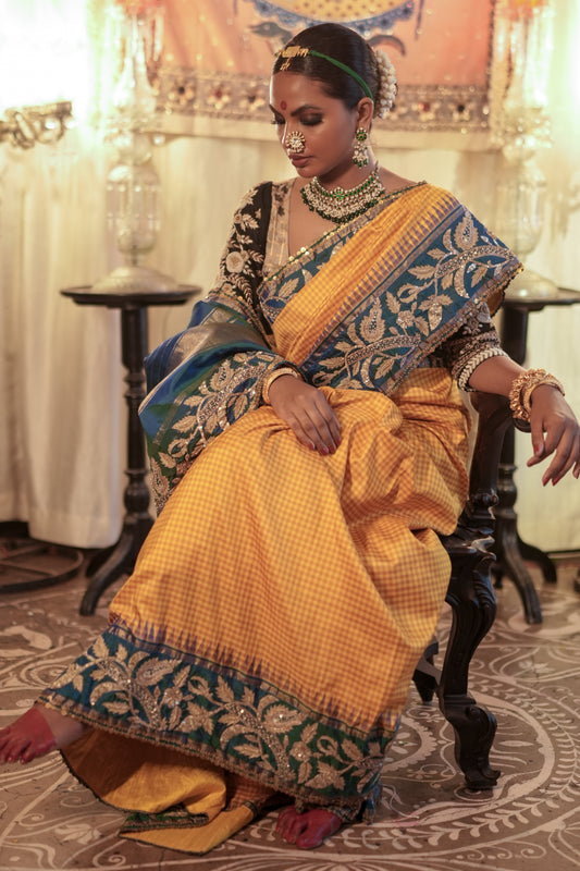 Woman in traditional yellow and blue kanjiveeram silk saree sitting on a patterned floor.