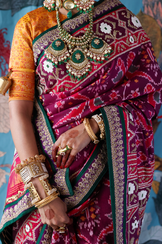Person wearing a colorful hand embroidered patola silk saree with intricate patterns and jewelry, holding a phone.