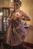 Woman in traditional handloom benarsi silk saree with intricate patterns and jewelry in a warm-toned room.